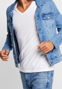 Man in een lichtblauwe denimjas over een witte V-hals T-shirt en bijpassende lichtblauwe jeans, halverwege een beweging tegen een effen achtergrond.