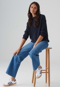 Marineblaue gerippte Strickjacke, hellblaue weit geschnittene Jeans und weiße Sneakers mit marineblauen Streifen. Holzsitz und hellgrauer Hintergrund.