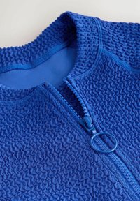 Mėlyna striukė su užtrauktuku ir tekstūruotu, susisukusiu audiniu. Turi apvalų užtrauktuko traukiklį ir kontrastingą lygią vidinę pamušalą.