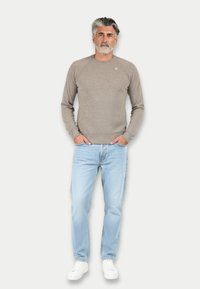 Maglione a maglia marrone chiaro con collo rotondo, abbinato a jeans azzurri e sneakers bianche. Il modello è in piedi con le mani nelle tasche.