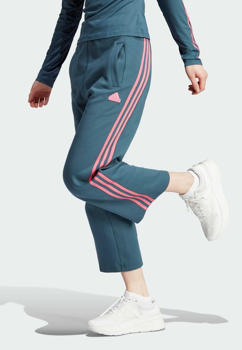 Tyrkysové zkrácené tepláky mají po stranách růžové tři pruhy, boční kapsy a logo Adidas na levém boku. Nošeny s bílými teniskami.