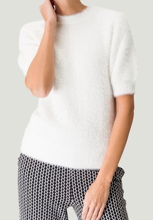 Persona que lleva un suéter blanco y esponjoso de manga corta y pantalones con un patrón en blanco y negro, sobre un fondo claro y liso.