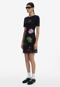 Schwarzes Kurzarmkleid mit bunten Blumenmustern, durchscheinendem unteren Teil, kombiniert mit schwarzen Sneakers und weißen Socken.