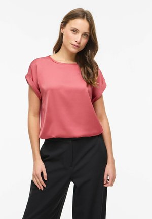 Žena s dlhými hnedými vlasmi, oblečená v krátkoslávkovom ružovom saténovom blúzke a čiernych nohaviciach s vysokým pásom, stojaca pred jednotným bielym pozadím.