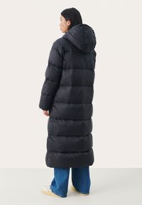 Lange zwarte pufferjas met een capuchon, met gewatteerde delen; gecombineerd met blauwe spijkerbroek en lichte sneakers.