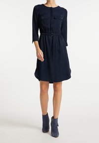 Vestido azul marino con mangas largas, dos bolsillos en el pecho, una cintura ajustada y una textura sutil, combinado con botas de tobillo azules.