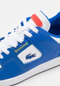 Baskets en cuir bleu avec accents blancs, comprenant un panneau en cuir blanc et un logo crocodile brodé. Comprend un marquage orange vif et des lacets blancs.