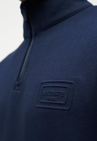 Personne portant un pull à quart de fermeture éclair bleu marine avec un patch texturé du logo Lacoste Paris sur la poitrine.