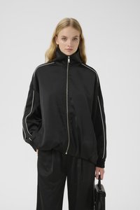Chaqueta negra de satén con cremallera, cuello alto, rayas laterales blancas, puños elásticos y un corte relajado, combinada con pantalones negros a juego.