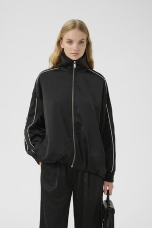 Black satin zip-up jacket with high collar, white side stripes, elastic cuffs, and relaxed fit, paired with matching black trousers.