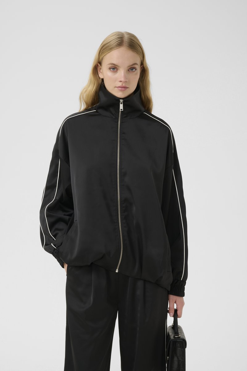 Chaqueta negra de satén con cremallera, cuello alto, rayas laterales blancas, puños elásticos y un corte relajado, combinada con pantalones negros a juego.