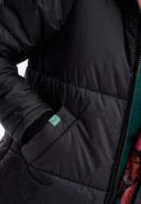 Čierna páperová bunda s prešívanou štruktúrou, so zipsom a bočnou kapsou s malou zelenou nášivkou. Podšívka je viditeľná s kvetinovým vzorom.