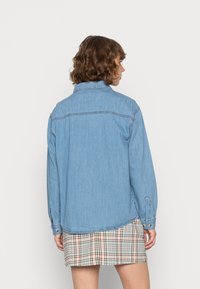 Camisa de ganga de mangas longas, cor azul clara, corte reto, punhos com botões e detalhes de costura visíveis. Usada sobre uma saia xadrez.