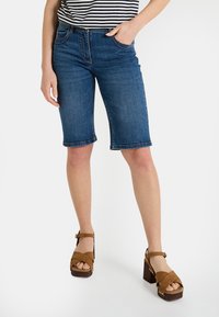 Shorts en denim bleu foncé, avec une coupe ajustée, un ourlet roulé et des poches latérales. Associés à des sandales compensées marron.
