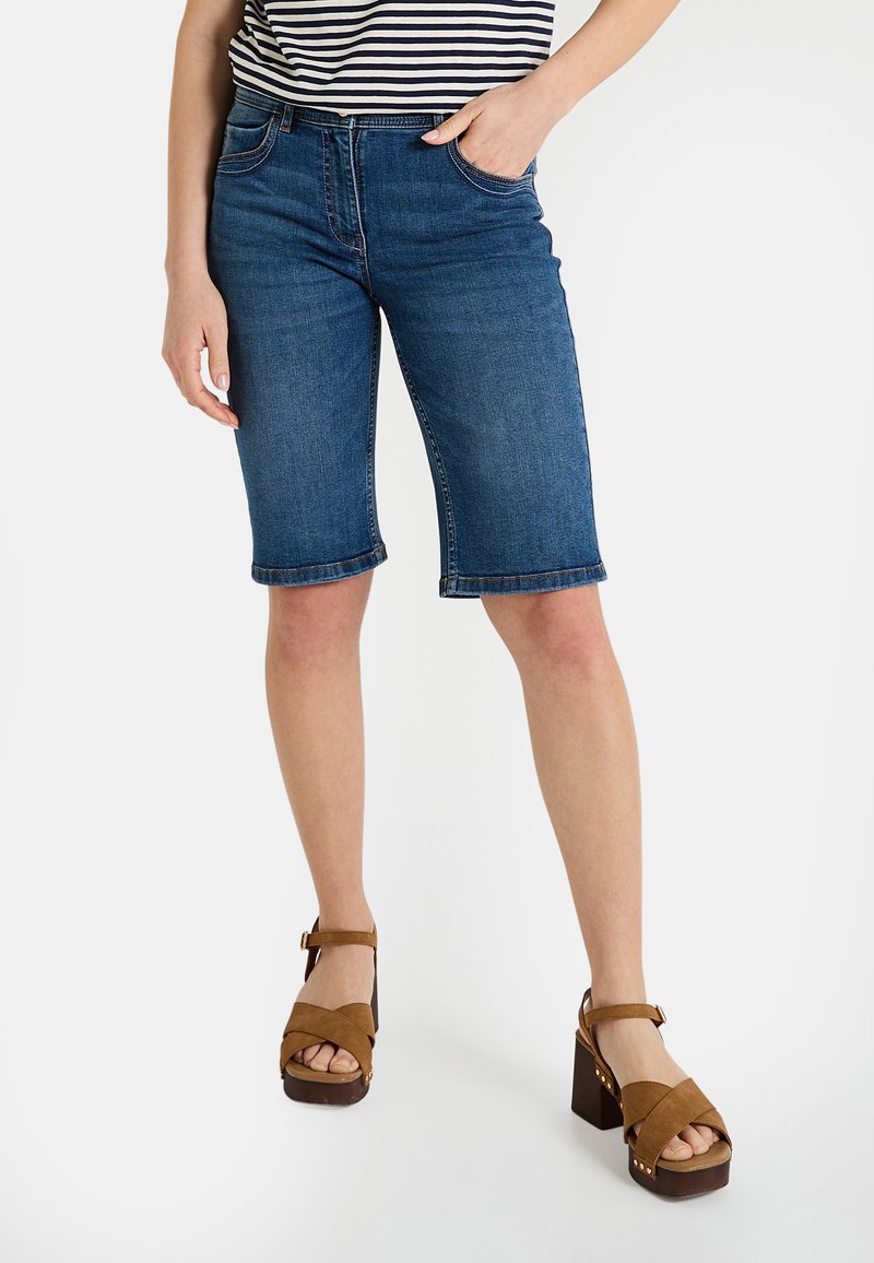Shorts en denim bleu foncé, avec une coupe ajustée, un ourlet roulé et des poches latérales. Associés à des sandales compensées marron.