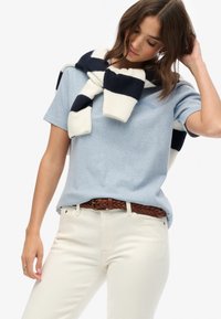 Lichtblauw katoenen T-shirt met korte mouwen, gecombineerd met crèmekleurige jeans en een marineblauw-wit gestreepte gebreide sjaal over de schouders gedrapeerd.