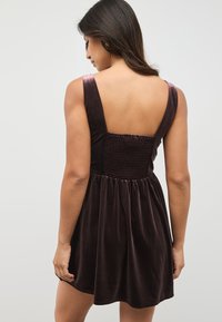 Žametna obleka v globoki vijolični barvi s prilegajočim se nazaj nabranim delom, širokimi naramnicami in razširjeno krilo do kolen. Gladek tekstura in eleganten dizajn.