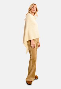 Poncho in lana beige con una texture morbida, caratterizzato da un design drappeggiato e bordi frangiati, abbinato a pantaloni in corduroy color nocciola e calzature marroni.