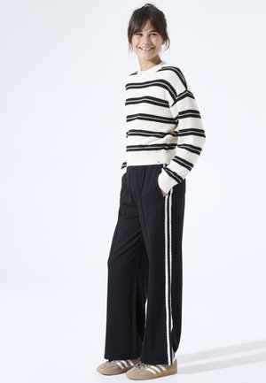 Young person smiling, wearing a white and black striped sweater, black wide-leg pants with white side stripes, and beige sneakers.