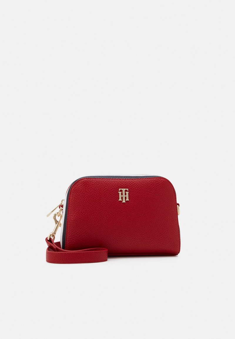 Punainen teksturoitu nahkainen käsilaukku, jossa on kultasävyinen logo ja vetoketju, varustettuna irrotettavalla hihnalla valkoisella taustalla.