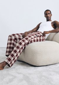 Pantalones de cuadros de algodón en burdeos y crema con una cintura elástica, combinados con una camiseta sin mangas blanca, usados mientras se está sentado en una silla de textura crema.