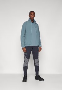 Veste zippée bleu clair avec col montant, superposée sur une chemise bleu foncé. Pantalons noirs avec accents et motifs gris, accompagnés de chaussures noires.
