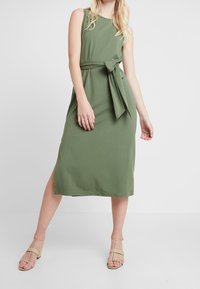 Abito verde senza maniche con vita cintata e dettaglio di nodo. Realizzato in tessuto liscio, presenta un taglio di lunghezza media e fessure laterali.