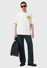 Hvid kortærmet t-shirt med en gul grafisk lomme, parret med mørkegrønne brede bukser og en sort lædertaske. Sorte sko og solbriller.
