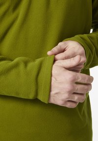 Zielona polarowa bluzka z długim rękawem i ściągaczami, ukazująca zbliżenie rąk dostosowujących ściągacz. Widać miękką teksturę i minimalne szwy.