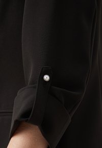 Blouse noire à la texture lisse, avec un poignet roulé maintenu par un bouton orné d'une perle et un design épuré.