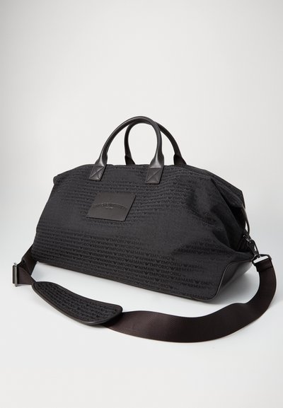 Sac de sport en tissu noir avec un motif de logo en relief, des poignées en cuir et une bandoulière réglable. Il possède une base marron solide et un design épuré.