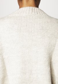 Sweter dzianinowy z wysokim, prążkowanym kołnierzem, wykonany z miękkiej, teksturowanej, kremowej przędzy. Charakteryzuje się luźnym krojem i widocznymi detalami przeszyć.