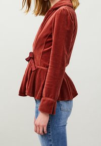 Chaqueta peplum de terciopelo oxido con manga larga, detalle de lazo en la cintura y puños ligeramente acampanados. Tejido texturizado; vista lateral. Jeans de denim debajo.