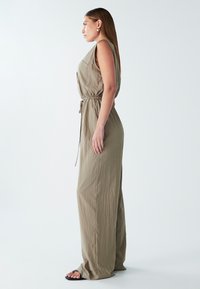 Khaki kombinéza s volným střihem, bez rukávů, s nastavitelným šňůrkovým pasem a bočními kapsami. Lehčí, texturovaná látka.