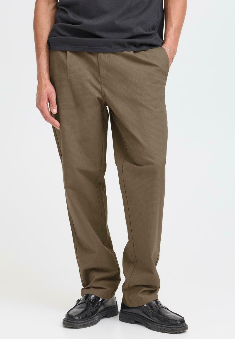 Una persona está de pie vestida con una camiseta negra y pantalones marrones, combinados con zapatos negros, sobre un fondo claro. Manos en los bolsillos.