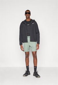 Chaqueta ligera negra con cremallera, camiseta negra ajustada, pantalones cortos verdes claros, calcetines negros y zapatillas deportivas negras con acentos de malla.