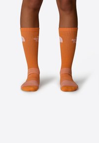 Medias naranjas hasta la rodilla con textura de costillas verticales y acentos rosas en los dedos y el puño. Presenta el logo de The North Face en blanco.
