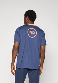 Men's navy athletic shirt featuring a coral accent on the collar, "BARÇA" logo in a circular design on the back, and short sleeves.