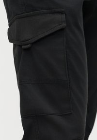 Primo piano della gamba di pantaloni cargo neri con tasca a patta e chiusura a bottone sul lato.