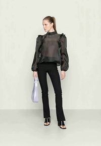 Blusa preta transparente com mangas bufantes, combinada com calças pretas ajustadas. A modelo segura uma bolsa lilás claro e usa sandálias de salto pretas.