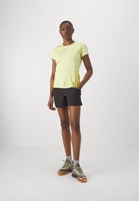 Camisa deportiva de manga corta, amarilla brillante, con una textura suave, combinada con pantalones cortos negros y zapatillas de trail resistentes en verde y naranja.