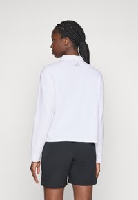 Vrouw met gevlochten haar, gekleed in een witte lange mouwen polo en zwarte shorts, van zich af kijkend tegen een effen lichte achtergrond.