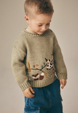 Lille barn iført en beige strikket sweater med dyre- og naturmotiver, kigger ned, stående mod en ensfarvet lys baggrund.