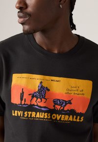 Nærbilde av en svart T-skjorte med et fargerikt vintage trykk av Levi Strauss overaller, som viser en cowboy på hesteryggen og en okse.