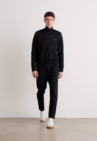 Costum de trening negru cu fermoar și guler înalt, având un design elegant și un logo subtil. Purtați cu adidași albi pe un fond neutru.
