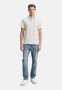 Polo de color beige claro con un patrón sutil, de manga corta y tres botones. Combinado con unos vaqueros azul claro y zapatillas blancas.