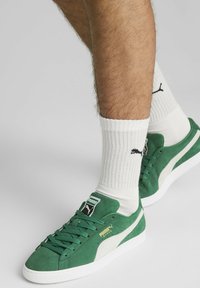 Zielone zamszowe sneakersy z białymi akcentami i teksturowaną podeszwą, z metką z logo i płaskimi sznurówkami. Noszone z białymi skarpetkami z prążkami.
