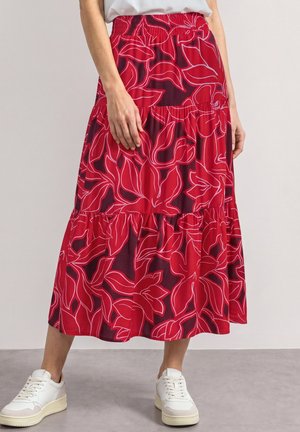 Person iført et rødt blomstrende lagdelt skørt med hvide og lyserøde mønstre, hvid t-shirt og hvide sneakers, står på et gråt gulv.