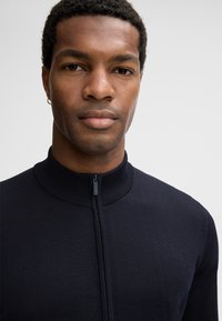 Ciemnogranatowy sweter na zamek z wysokim kołnierzem. Dzianinowa faktura, dopasowany krój widoczny wokół szyi i klatki piersiowej.