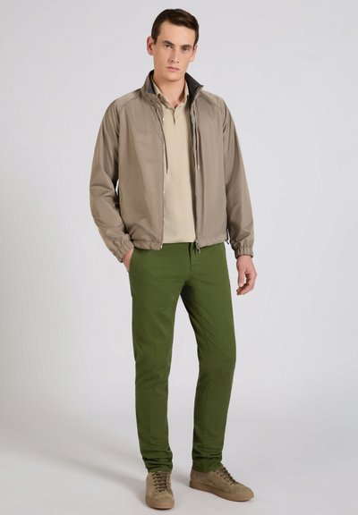 Man wearing beige zip-up jacket, light beige shirt, olive green pants, and beige sneakers standing with hands partly in pockets.